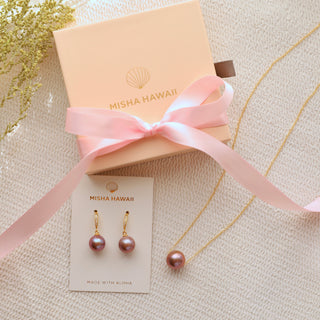 A pink pearl necklace and earrings set with a gold-filled and vermeil finish, presented next to a pink box with 'misha hawaii' branding, on a white textured surface with a decorative branch in the background.