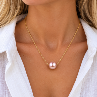A close-up of a person wearing a delicate gold chain necklace with a single round, lustrous pink pearl pendant, styled with a white collared shirt.