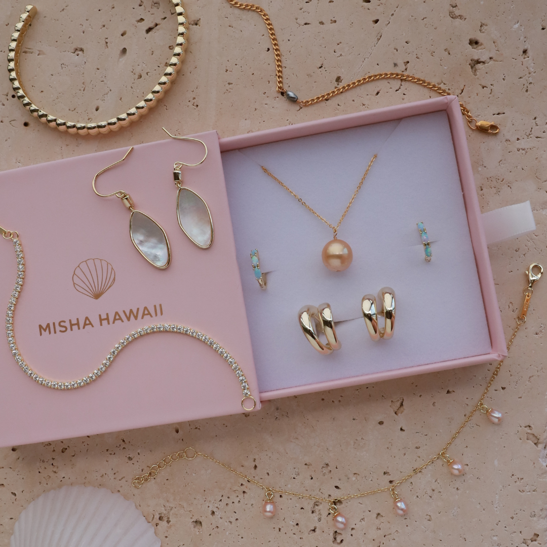 A jewelry gift set from Misha Hawaii displayed in an open pink box on a beige stone surface, with additional gold jewelry and a seashell arranged around it. The set includes a pearl necklace, gold hoop earrings, drop earrings with iridescent inlays, small blue and pearl stud earrings, and a beaded gold bracelet.