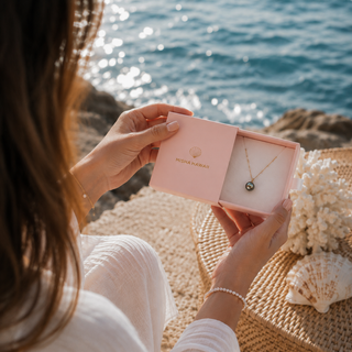 A person sitting by the ocean holds an open pink jewelry box displaying a delicate gold necklace with a small round pendant. The box is branded with the name Misha Hawaii