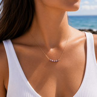 A close-up lifestyle shot of a woman wearing a delicate gold necklace with five small white pearls on a beach with the ocean in the background.