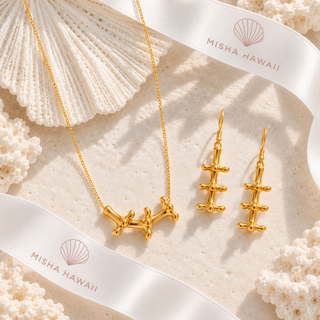 Gold jewelry set featuring a necklace and matching drop earrings with a coral-like barbell design, styled on a textured beige surface with white seashell props and white ribbons printed with the MISHA HAWAII brand name.