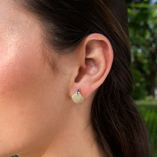 Close-up of an ear wearing a gold shell earring with a blue gemstone, blurred green background