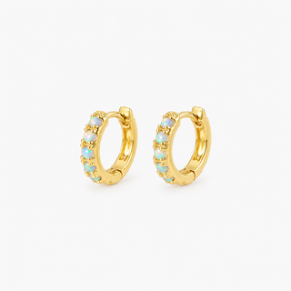 A pair of gold huggie hoop earrings with small light blue opal-like stones set along the front, shown on a clean white background.