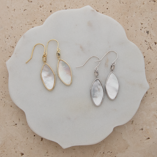 Two pairs of teardrop-shaped dangle earrings displayed on a scalloped white stone tray. One pair has gold-tone metal settings with iridescent white mother-of-pearl drops, and the other has silver-tone metal settings with matching iridescent white drops.