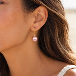 A close-up of a woman's ear wearing a gold huggie hoop earring with a single dangling pink freshwater pearl.