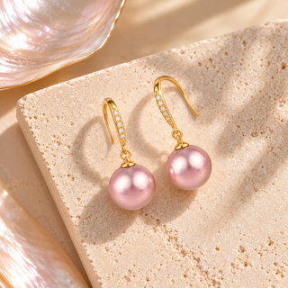 A pair of gold drop earrings with large pink freshwater pearls and small clear stones along the hooks, displayed on a textured beige stone slab.