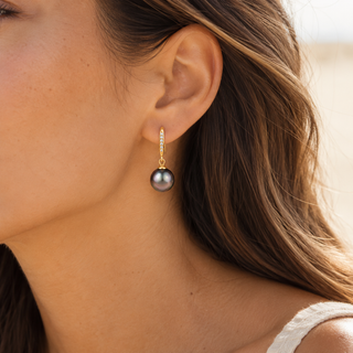 A close-up of a woman's ear wearing a gold drop earring with a single dark gray pearl and a small row of clear stones above it.