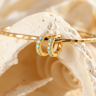 Gold huggie hoop earrings with light blue stones, displayed on a gold bangle over a white seashell prop.