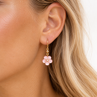 A close-up of a woman's ear wearing a dangling earring with a small pink flower charm and gold-tone hooks.