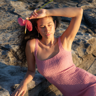 Model wearing a pink dress adorned with pink keshi pearl strand, holding a flower while relaxing on rocks.