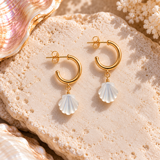 A pair of gold hoop earrings with white shell-shaped drop charms, displayed on a textured beige stone with seashells and coral props.