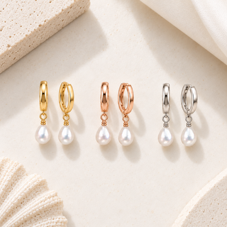 three pairs of hoop dangle earrings arranged in a row on a light beige textured surface. The earrings come in yellow gold, rose gold, and silver metal finishes, each with a small teardrop-shaped white pearl drop.
