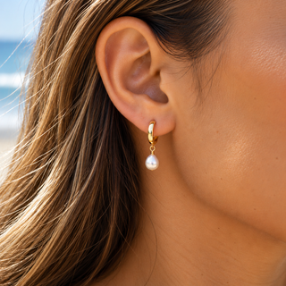 A close-up of a woman's ear wearing a small gold huggie hoop earring with a dangling white pearl drop, with her hair and skin visible against a softly blurred beach background.