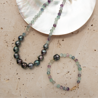 A beaded necklace and matching bracelet displayed on a textured beige surface with a large iridescent shell disc. The jewelry features faceted beads in pale mint green, lavender purple, and dark iridescent gray-green with a small gold clasp.