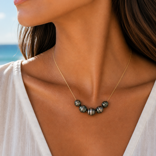 A close-up lifestyle shot of a woman wearing a delicate gold chain necklace with a row of dark gray and metallic silver beads at the center, set against a soft-focus beach background.