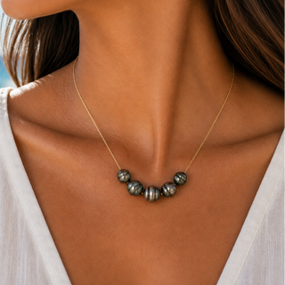 A close-up lifestyle shot of a woman wearing a delicate gold chain necklace with a row of peacock pearls at the center, set against a soft-focus beach background.