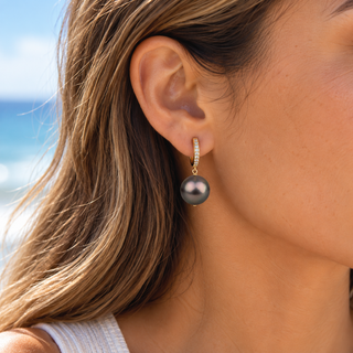 A close-up of a woman's ear wearing a gold huggie hoop earring with a small diamond-set band and a dangling dark gray freshwater pearl, with a blurred beach background.