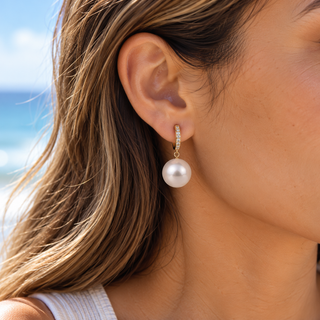 A close-up of a woman's ear wearing a single drop earring with a small gold huggie setting and a lustrous white pearl pendant, photographed outdoors with a blurred beach background.
