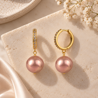 A pair of gold hoop earrings with a pavé-set clear stone band and a dangling pink pearl, displayed on a textured cream stone slab with decorative white flowers and satin fabric.
