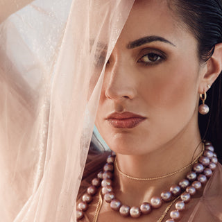A close-up portrait of a woman wearing layered pearl necklaces, a gold chain necklace, and a pearl drop earring, with a sheer light pink veil draped over her face.