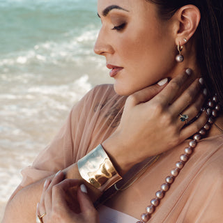 A woman in a peach sheer top poses by the ocean, showcasing a gold cuff bracelet, pearl drop earrings, and a pearl necklace.