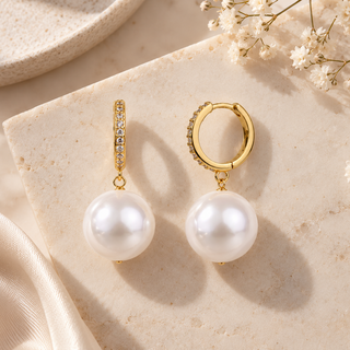 A pair of gold hoop and drop earrings displayed on a beige stone slab with dried flowers and satin fabric. The earrings feature white freshwater pearls and small clear stones set in gold.