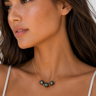 A close-up of a woman wearing a delicate gold necklace with three dark, iridescent baroque pearls at the center, styled with a white spaghetti strap top.