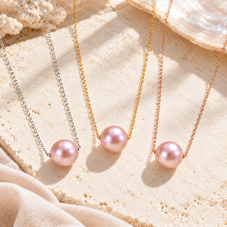 Three pearl necklaces with delicate chains in silver, gold, and rose gold, displayed on a textured beige stone surface with a seashell prop.