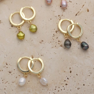 Four pairs of gold hoop earrings with dangling pearl-like beads in olive green, dark gray, white, and light pink, arranged on a textured beige surface with a white fabric prop.