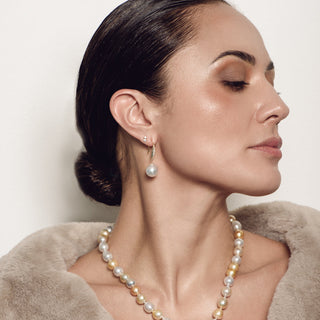 A woman in profile wearing a pearl necklace and matching drop earrings, with dark hair in a low bun and a beige faux-fur collar against a plain light background.