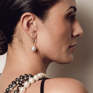 A close-up side profile of a woman wearing a stack of pearl necklaces and a single pearl drop earring with a gold huggie hoop.