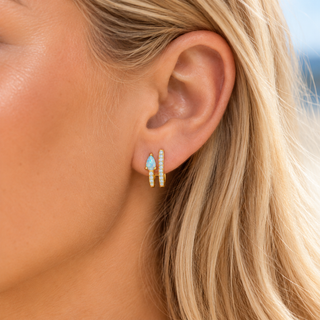 A close-up of a woman's ear wearing a pair of small gold huggie hoop earrings with light blue teardrop-shaped stones and clear crystal accents.