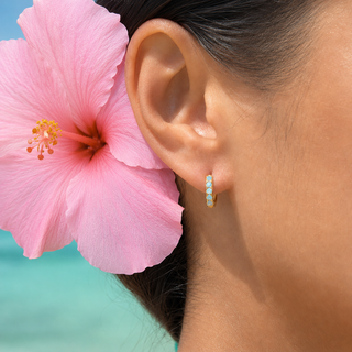 A close-up side profile of a woman's ear wearing a small gold huggie hoop earring set with light blue stones, with a large pink hibiscus flower tucked behind her ear and a turquoise background.