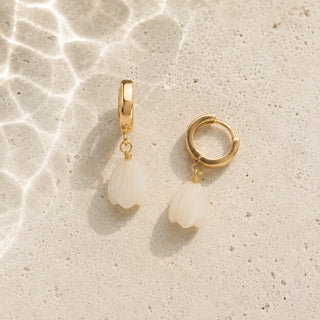 A pair of gold huggie hoop earrings with dangling white shell-shaped charms, displayed on a textured beige stone surface with water ripple shadows.
