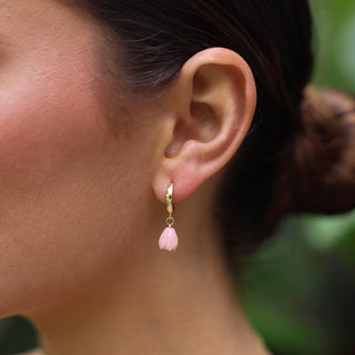A close-up of a person's ear wearing a small gold huggie hoop earring with a dangling pink flower charm.