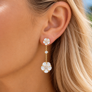 A close-up of a model's ear wearing a gold dangle earring with two white mother-of-pearl flower charms and a small clear rhinestone in the middle.
