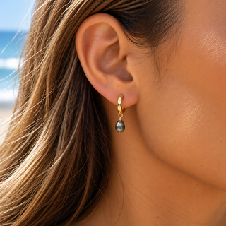 A close-up of a woman's ear wearing a small gold huggie hoop earring with a dangling gray labradorite stone, set against a blurred beach background.