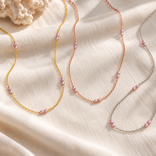 Three delicate necklace chains laid out on cream fabric: one gold-tone, one rose gold-tone, and one silver-tone, each accented with small pink freshwater pearls. A coral fragment appears in the top-left corner.