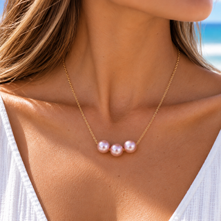 A close-up of a woman's neck and chest wearing a delicate gold chain necklace with three small, lustrous pink pearls at the center, set against a softly blurred beach background.