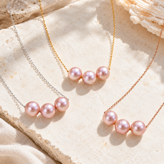 Three pearl bar necklaces with pink freshwater pearls on delicate chains, styled on a textured beige stone surface with a seashell prop.