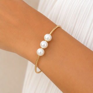 A delicate gold wire bracelet with three white faux pearls, shown worn on a person's wrist against a white pleated top.