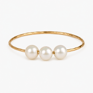 A gold bangle bracelet featuring three smooth, lustrous white pearls aligned along the front, set against a clean white background.