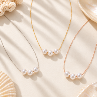 Three pearl bead necklaces with delicate chains are arranged on a light beige fabric background with soft shadows and decorative seashell props. The necklaces feature white pearls set in silver, gold, and rose gold tones.