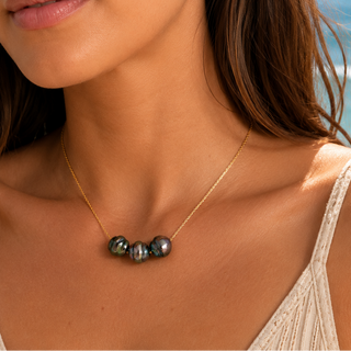 A close-up lifestyle shot of a woman wearing a delicate gold chain necklace with four dark, iridescent baroque pearls at the center, set against a blurred beach background with blue ocean water.