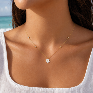 A close-up lifestyle shot of a woman wearing a delicate gold necklace with a small white flower pendant and tiny gold bead accents, set against a blurred beach background.
