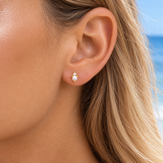A close-up of a woman's ear wearing a small gold stud earring with a white pearl, set against a blurred beach background.